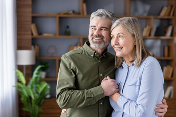 Senior couple embracing and smiling as they look out a window together in their cozy living room, radiating love, companionship, contentment and hopeful retirement life