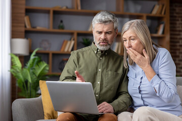 Senior couple sitting on a sofa at home, looking at a laptop screen with expressions of shock and worry after discovering unexpected bad news, a scam, or a high bill online