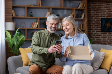 Senior couple sitting on a sofa, looking at a smartphone screen, happy about their new online purchase, receiving a package, sharing a moment of excitement and connection with digital technology