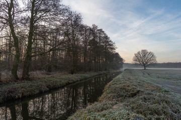 Winter canal with bare trees and frosty fields at sunrise