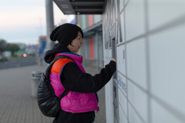 Woman picking parcel from locker kiosk, soft evening light, backpacked commuter taps touchscreen to...