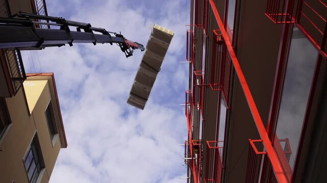 A construction crane lifts insulated roofing panels. A hydraulic truck crane precisely moves large, shrink-wrapped stacks of insulated sandwich panels between closely spaced residential buildings.