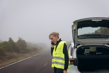 Woman in reflective vest standing by car on foggy road, experiencing vehicle trouble and seeking assistance