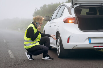 Woman in reflective vest kneeling beside car on roadside, experiencing vehicle trouble while making a phone call for assistance