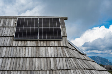 Modern solar panels installed on the traditional shingled wooden roof of a shepherd's hut against a cloudy sky in Slovenia