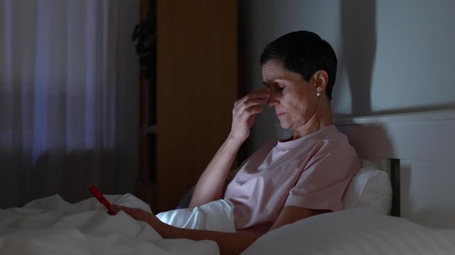 Tired senior woman wasting time in bed scrolling social media on smartphone late at night and feeling unwell. Mature lady with insomnia and screen addiction procrastinating late hours with phone.