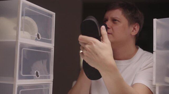 Man sniffing black sandals for odor before organizing shoe collection in transparent plastic containers, ensuring clean storage and preventing unpleasant smell for seasonal cupboard space arrangement.