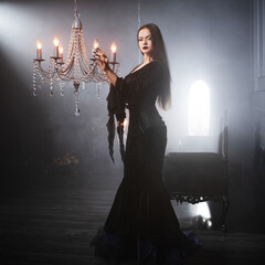 A gothic femme fatale with long straight hair and dark makeup wears a black godet skirt dress with corset, posed in a luxurious dimly lit gothic interior. © Ulia Koltyrina
