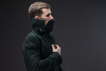 Male model in warm coat with tall collar, depressive cyberpunk fashion on gray.