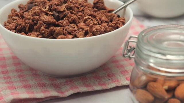 Bowl of chocolate granola breakfast on table with glass of milk, fruits and nuts, slow motion