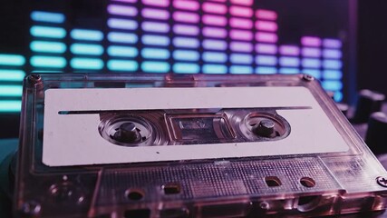 A closeup shot of a vintage transparent audio cassette tape resting in the foreground with a vibrant blurred digital sound equalizer displaying colorful pink blue and purple bars in the background ev.