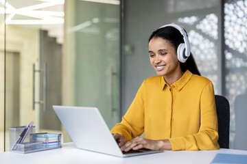 Young woman wearing white headphones smiling while confidently typing on a laptop, engaging in remote work or online meeting from a modern office environment