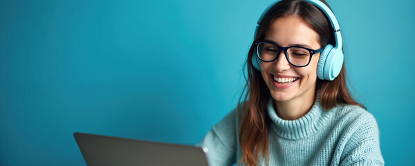 Young woman wears headphones and glasses, smiles at laptop screen, receiving positive remote learning message. She studies online, works from home, enjoys digital connection and modern education.