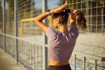 Woman athlete prepares ponytail for fitness training. Outdoors by a metal fence near sand court, she adjusts hair and tracker, focused and calm. Concept of active lifestyle and wellness.