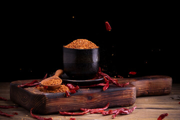 Chili powder in black bowl and wooden spoon with garlic, shallots, spicy spice mix. Asian food ingredients arranged on wooden table. Scattered chili powder background.