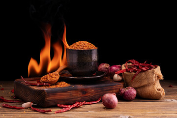 Chili powder in black bowl and wooden spoon with garlic, shallots, garden spicy spice mix. Asian food ingredients arranged on wooden table. Flame background, spicy food concept.