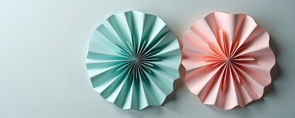 Two pastel paper rosettes in soft pink and teal shades adorn a plain wall. These handmade fans offer a cheerful, minimalist decoration for celebrations and events.