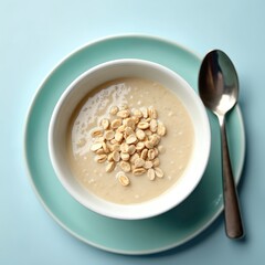 White bowl with creamy oatmeal porridge topped with rolled oats. A metal spoon rests on a light blue plate next to the dish. This meal is served on a solid pastel blue surface, ready for breakfast.