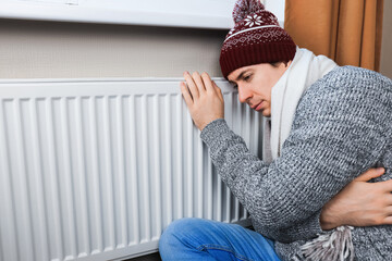 Freezing man in winter hat touching cold radiator at home. Heating trouble and energy crisis...