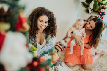 Happy diverse family with a baby decorating a Christmas tree in a cozy living room, celebrating the winter holidays together at home.