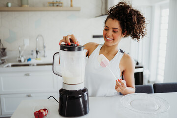 Happy young woman preparing a healthy smoothie using a blender in a bright, modern kitchen at home.