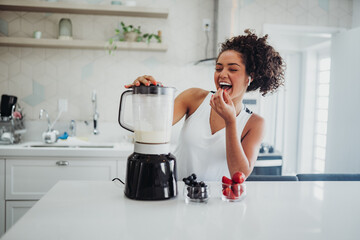 Joyful young woman with curly hair preparing a nutritious smoothie in a blender while snacking on fresh berries in a bright, modern kitchen.