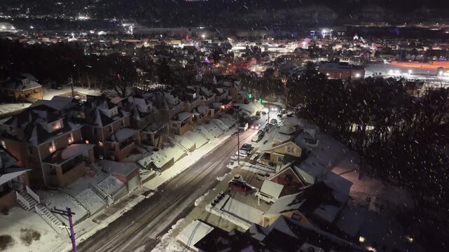 Snowy nighttime aerial view of a Pennsylvania residential neighborhood with glowing streetlights, moving cars, and falling snow creating a calm winter atmosphere. Pittsburgh suburbs. 15006  