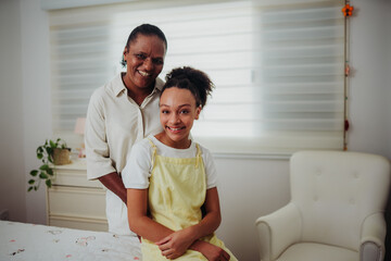 Happy African American mother and teenage daughter smiling together in a bright bedroom at home.