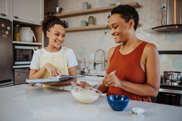 Happy African American mother and teenage daughter following a recipe from a cookbook while baking together in a modern kitchen.