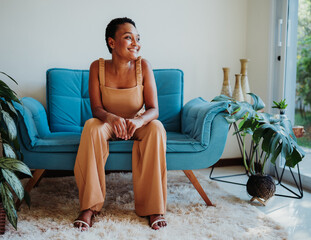 Happy African American woman sitting on a blue sofa in a modern, sunlit living room with indoor plants.