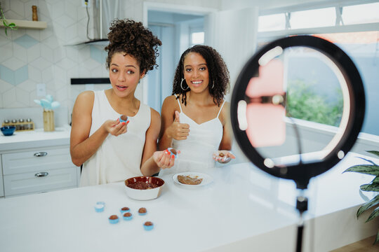 Two young Black female influencers recording a cooking tutorial for social media in a modern kitchen, using a ring light and smartphone to film chocolate truffle making.