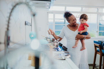 Smiling senior Black woman holding a cute baby while preparing food on a stove in a sunny domestic kitchen.