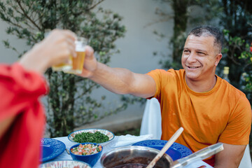 Smiling mature man toasting with a glass of beer during a traditional Brazilian lunch outdoors with friends.