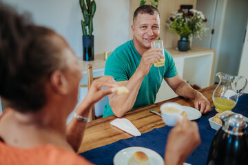 Happy middle-aged man drinking orange juice while enjoying breakfast with his partner at home.