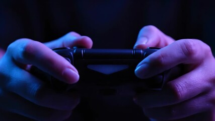Close-up of hands on a game controller in a dark room with vibrant blue and purple neon lighting.