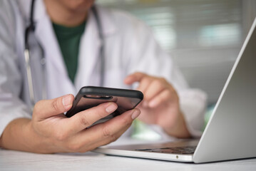 Doctor using smartphone and laptop for online consultation with patient. Physician texting on mobile phone in hospital office. Telemedicine, telehealth and digital healthcare technology concept.