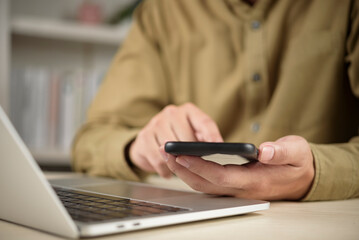 Close up of man hands holding smartphone and using laptop computer for online banking, business communication, and working from home concept.
