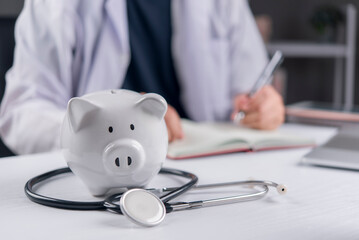 Piggy bank with stethoscope on doctor desk. Health insurance, medical cost and healthcare savings concept. Doctor writing medical prescription in background.
