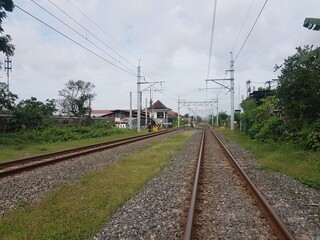 Railway tracks leading toward a small station building in the distance. Infrastructure and overhead power lines on a bright cloudy day. Train tracks and green nature leading to a destination.