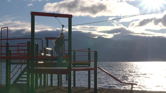 A young girl jumps off the platform at a playground on the beach, to ride the zipline. Wearing warm clothes on a mixed of clouds and sunny day, happy to be playing outside.