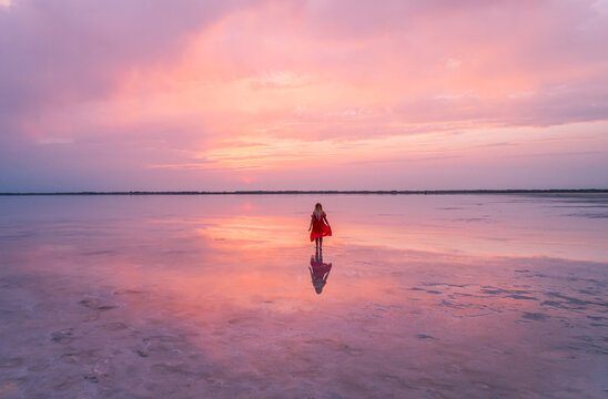 Aerial of a young woman in red dress walking in the water of a unique pink salt lake. Sunset at lake Bursol with beautiful reflections on calm water surface. Stunning scenery