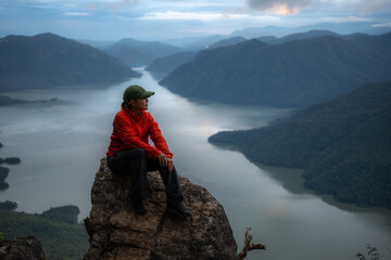 An Asian woman in a red dress sits on a rock on a mountain with other mountains and a river in the background.