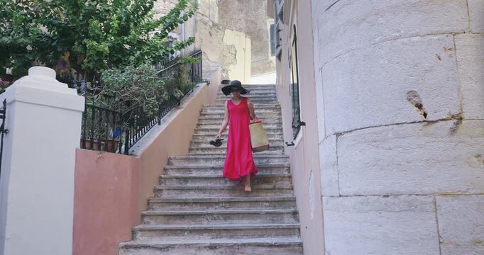 Woman tourist traveler in red dress and a hat walks on the street of the ancient Greek city, slow motion