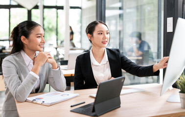 Two professional Asian businesswomen discussing financial reports and data analysis on a computer screen in a modern office, collaboration and teamwork concept.