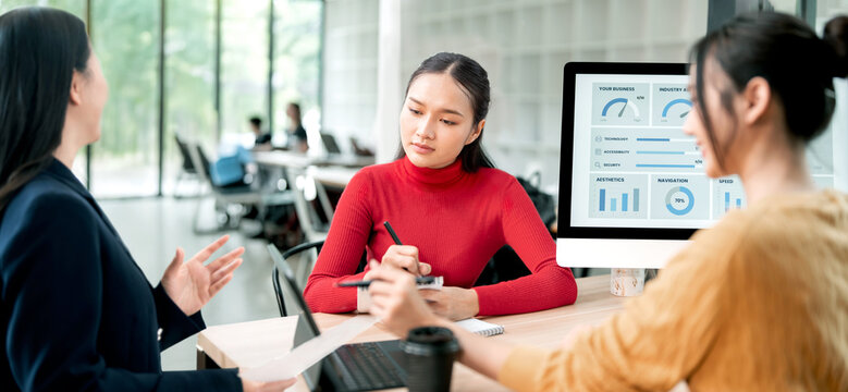 Diverse Asian businesswomen discussing financial charts and data on a monitor during a creative meeting in a modern office, teamwork concept. - Powered by Adobe
