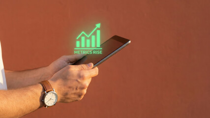High level planning concept with glowing metrics rise graph and confident hand holding digital tablet against warm urban wall background