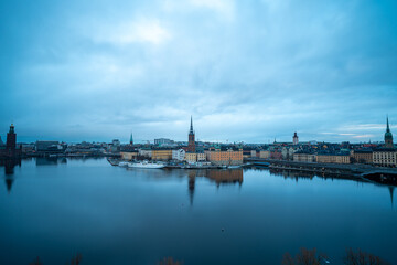 Stockholm skyline across Riddarfjärden with Riddarholmen Church and Stockholm City Hall reflected on calm water under an overcast sky, Sweden.