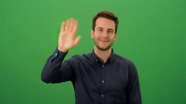 Confident young man with a friendly smile extends a warm greeting, waving his hand in a welcoming gesture against a clear green screen background, ideal for various media applications