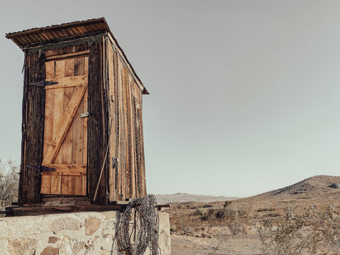 A small wooden outhouse with a slanted roof stands on a stone base under a clear sky surrounded by dry bushes and hills in a rustic remote outdoor scene