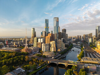 Naklejka premium Melbourne's expansive urban landscape featuring towering skyscrapers, the winding Yarra River, bridges, and green spaces bathed in golden light.